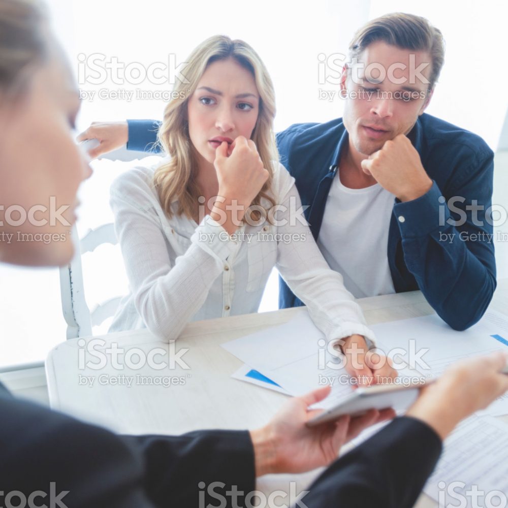 Real estate agent with couple looking through documents. The agent is holding a digital tablet showing it to the clients. Couple are casually dressed. They are looking concerned and worried and upset. Over the shoulder view.